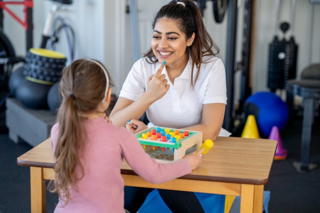 Speech therapist working with a girl by pointing on the mouth and smiling wide