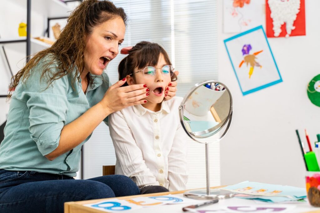 speech therapist working with a girl patient with mouth opening in front of the mirror